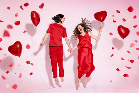 Playful couple celebrating Valentine’s Day, jumping hand in hand with red heart balloons and rose petals on a soft pink background.